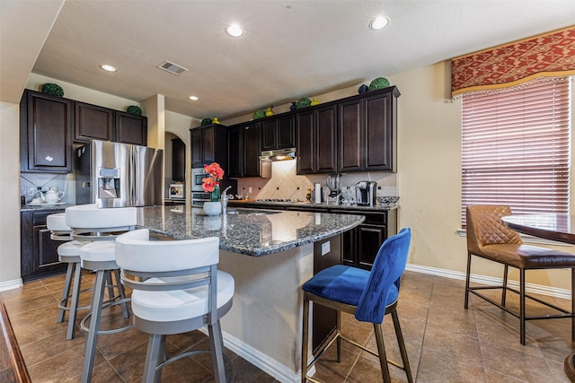 kitchen featuring tasteful backsplash, dark brown cabinets, stainless steel appliances, a center island with sink, and a breakfast bar area