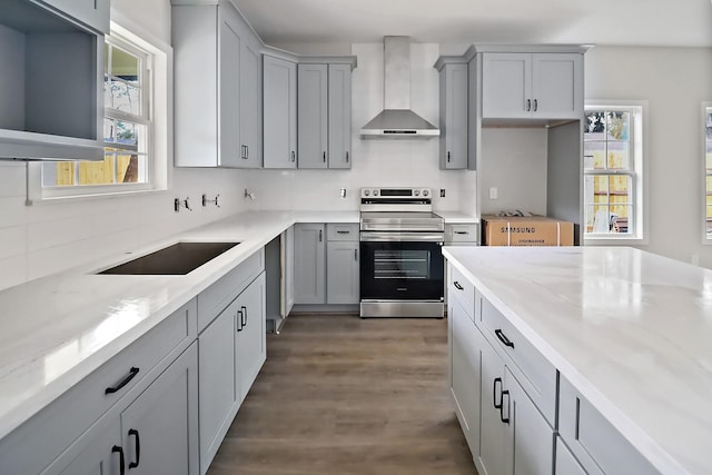 kitchen with wall chimney range hood, a wealth of natural light, light stone counters, and stainless steel range with electric stovetop