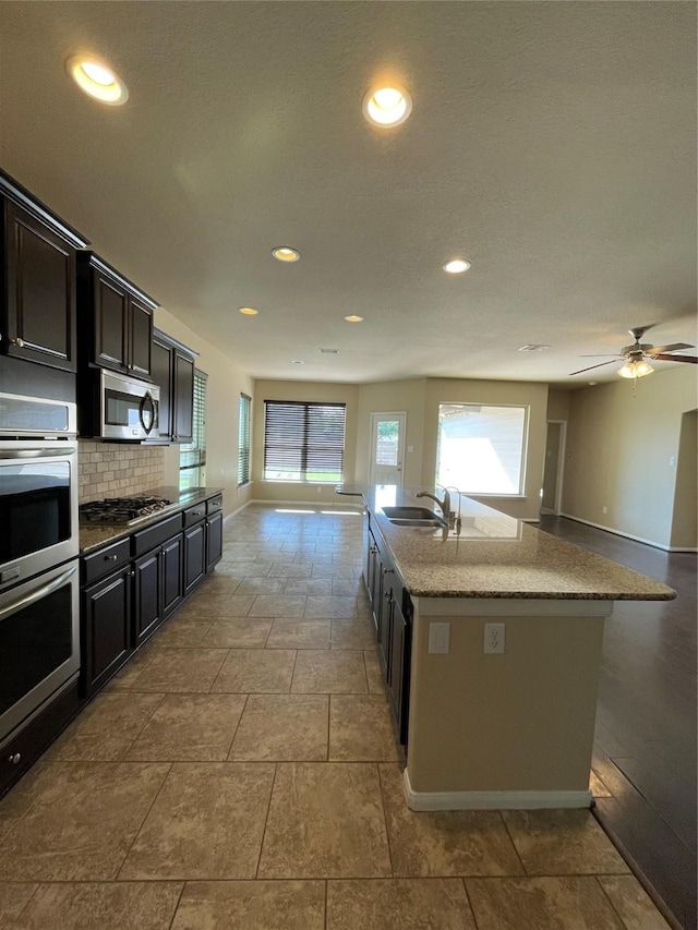 kitchen featuring tasteful backsplash, stainless steel appliances, ceiling fan, sink, and an island with sink