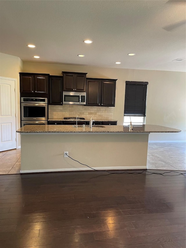 kitchen with stone counters, stainless steel appliances, tasteful backsplash, hardwood / wood-style floors, and a center island with sink