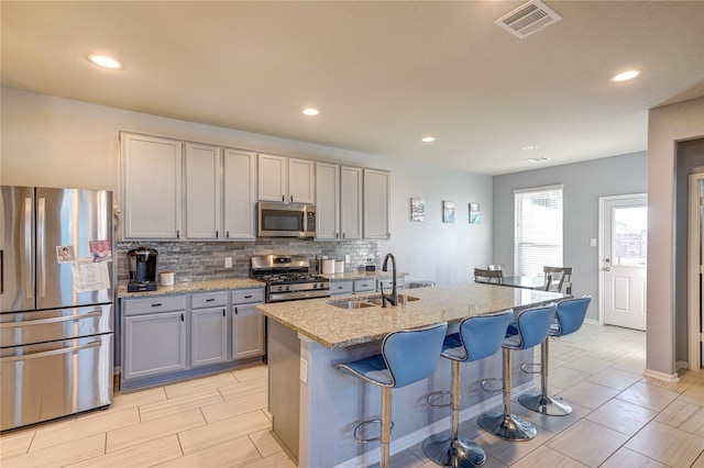 kitchen with gray cabinetry, sink, an island with sink, appliances with stainless steel finishes, and light stone counters