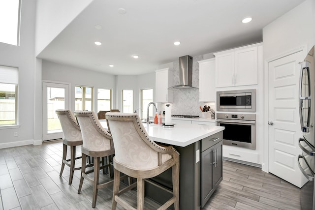 kitchen with white cabinets, wall chimney exhaust hood, an island with sink, a kitchen bar, and stainless steel appliances