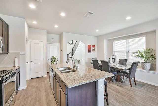 kitchen with stainless steel gas range oven, a kitchen island with sink, sink, light hardwood / wood-style flooring, and dark brown cabinets