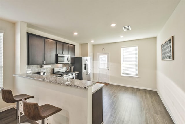 kitchen with light stone counters, stainless steel appliances, a peninsula, wood finished floors, and dark brown cabinets