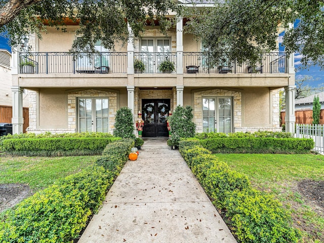 view of exterior entry featuring a balcony, fence, stone siding, french doors, and stucco siding