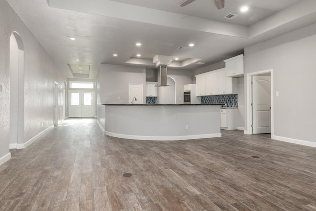 kitchen with wall chimney range hood, dark hardwood / wood-style flooring, an island with sink, a tray ceiling, and white cabinets