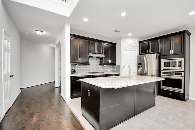 kitchen featuring backsplash, a kitchen island with sink, light stone countertops, appliances with stainless steel finishes, and light hardwood / wood-style floors