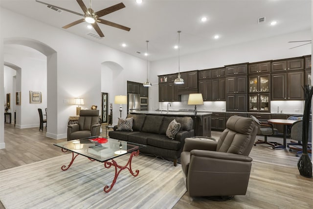 living room with ceiling fan, a towering ceiling, sink, and light hardwood / wood-style flooring