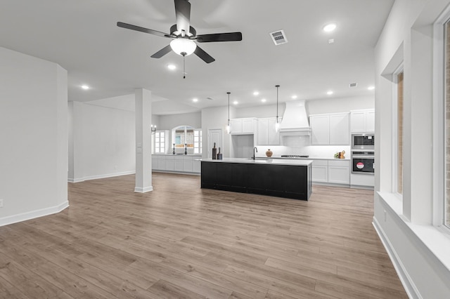 kitchen with decorative light fixtures, premium range hood, sink, white cabinetry, and an island with sink