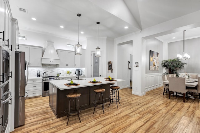 kitchen featuring appliances with stainless steel finishes, light wood-type flooring, custom range hood, a center island with sink, and hanging light fixtures