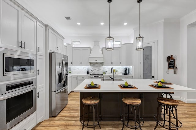 kitchen featuring a breakfast bar, custom exhaust hood, a kitchen island with sink, sink, and appliances with stainless steel finishes