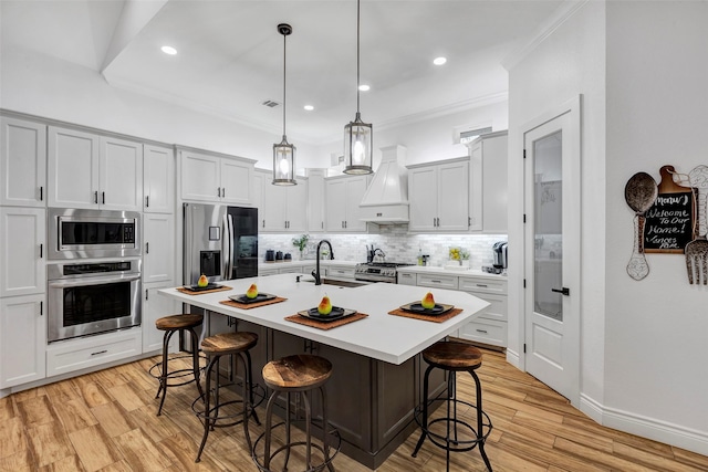 kitchen featuring appliances with stainless steel finishes, a kitchen breakfast bar, custom range hood, a center island with sink, and light hardwood / wood-style flooring