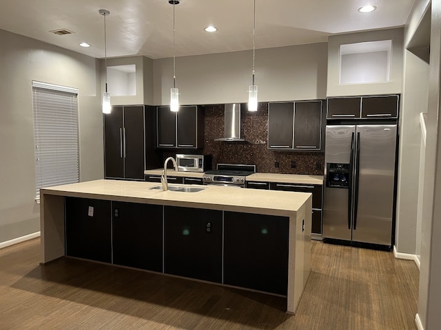 kitchen featuring appliances with stainless steel finishes, wall chimney exhaust hood, sink, hanging light fixtures, and an island with sink