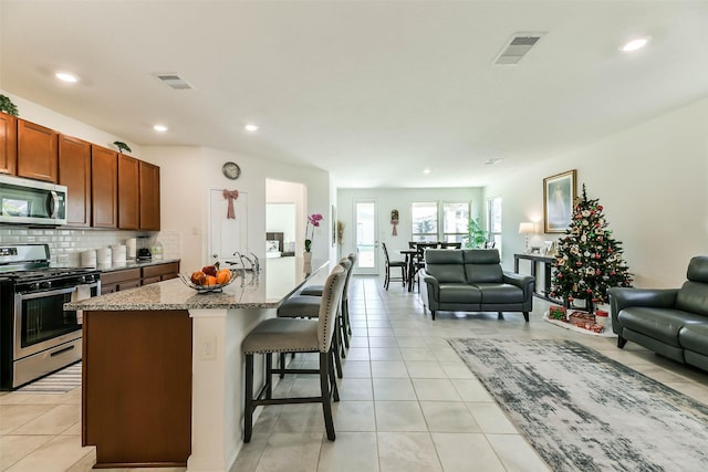 kitchen featuring a breakfast bar, a center island with sink, appliances with stainless steel finishes, light tile patterned flooring, and light stone counters