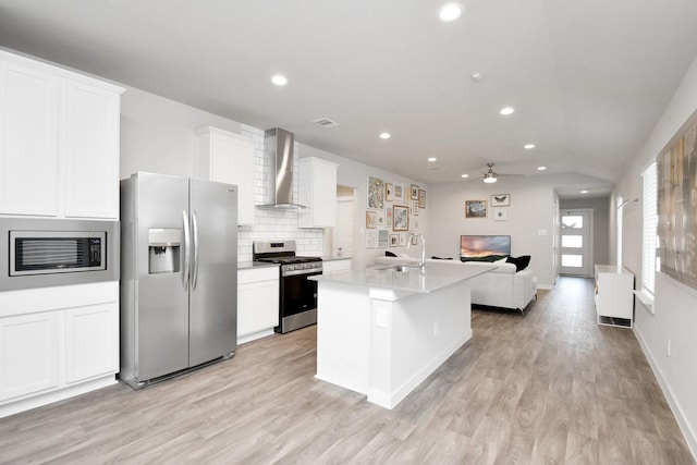 kitchen featuring wall chimney exhaust hood, stainless steel appliances, a kitchen island with sink, white cabinets, and light wood-type flooring