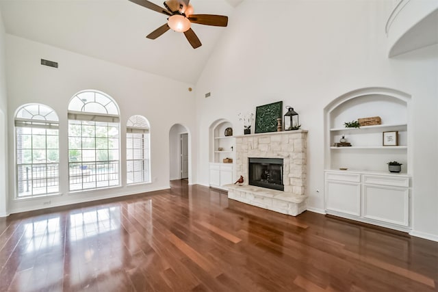 unfurnished living room with ceiling fan, dark wood-type flooring, high vaulted ceiling, built in features, and a stone fireplace