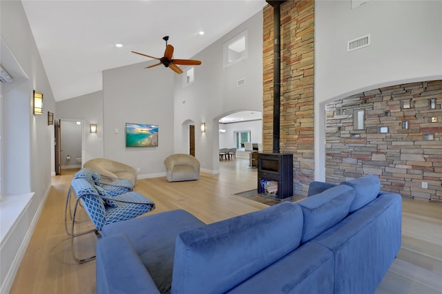 living room featuring ceiling fan, a wood stove, a towering ceiling, and light hardwood / wood-style flooring