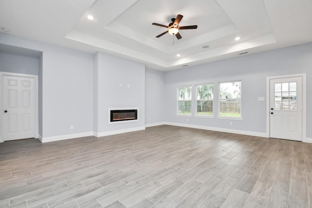 unfurnished living room featuring ceiling fan, a healthy amount of sunlight, a raised ceiling, and light hardwood / wood-style flooring