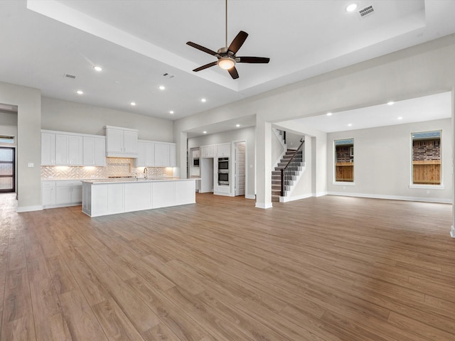 unfurnished living room with light wood-type flooring, a raised ceiling, ceiling fan, and sink