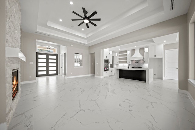 unfurnished living room featuring french doors, a tray ceiling, ceiling fan with notable chandelier, and a stone fireplace