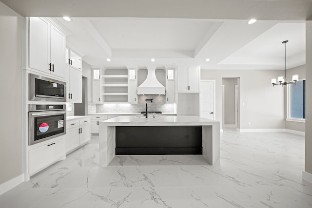 kitchen with premium range hood, a raised ceiling, stainless steel appliances, a kitchen island with sink, and white cabinets