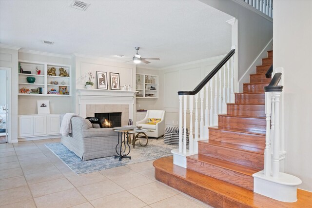 tiled living room with built in shelves, ceiling fan, a fireplace, and ornamental molding