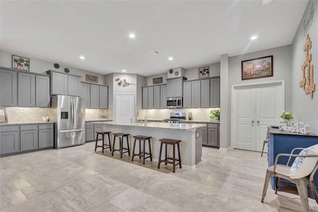 kitchen featuring stainless steel appliances, light stone counters, gray cabinets, a kitchen island with sink, and a breakfast bar