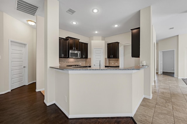 kitchen with backsplash, kitchen peninsula, tile patterned flooring, light stone countertops, and dark brown cabinetry