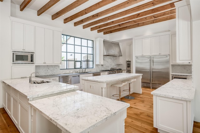 kitchen with a center island, wall chimney range hood, built in appliances, beam ceiling, and light stone counters