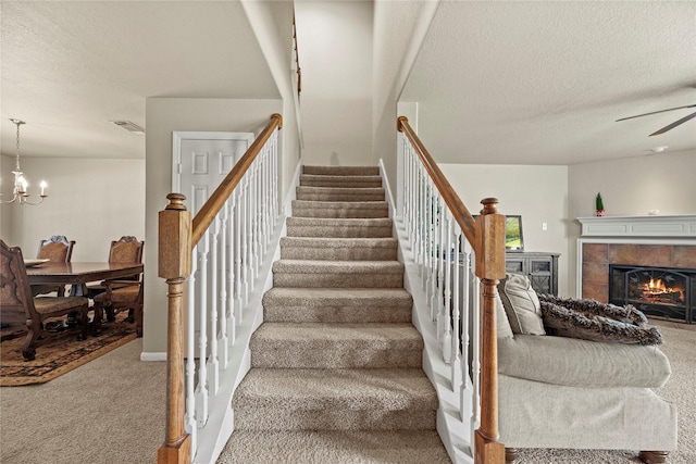 stairs featuring ceiling fan with notable chandelier, carpet floors, a textured ceiling, and a tile fireplace