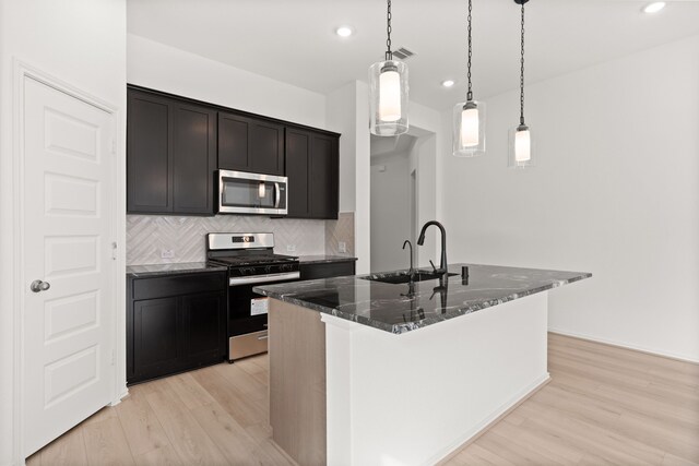 kitchen with sink, light hardwood / wood-style flooring, dark stone countertops, a kitchen island with sink, and appliances with stainless steel finishes