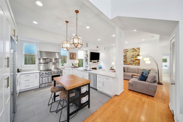 kitchen with wall chimney exhaust hood, hanging light fixtures, appliances with stainless steel finishes, white cabinets, and light wood-type flooring