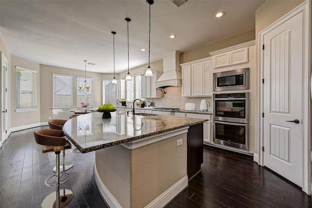 kitchen with light stone countertops, appliances with stainless steel finishes, custom exhaust hood, a center island with sink, and white cabinets