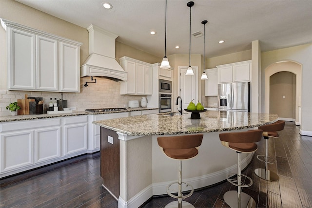 kitchen featuring white cabinetry, tasteful backsplash, premium range hood, a kitchen island with sink, and appliances with stainless steel finishes