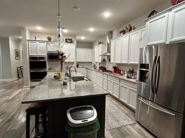 kitchen featuring a center island with sink, decorative light fixtures, wall chimney range hood, and appliances with stainless steel finishes