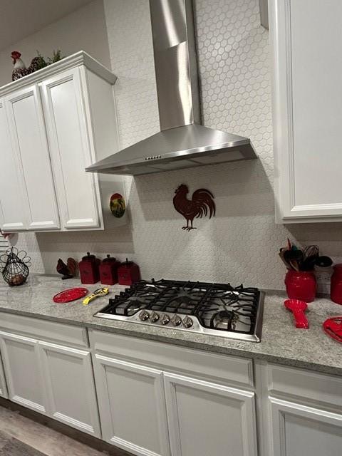 kitchen with wall chimney range hood, stainless steel gas stovetop, white cabinetry, and light stone countertops