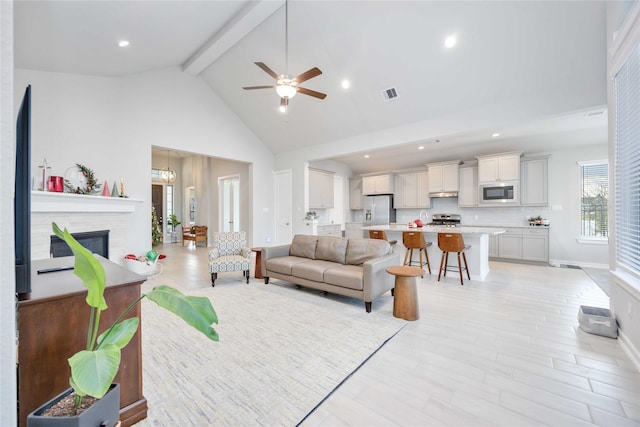 living room featuring high vaulted ceiling, ceiling fan with notable chandelier, a fireplace, beamed ceiling, and light hardwood / wood-style floors