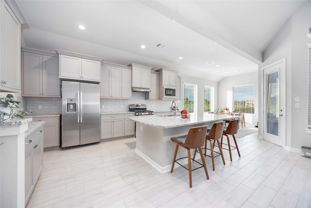 kitchen with a kitchen island with sink, vaulted ceiling, light stone countertops, appliances with stainless steel finishes, and a breakfast bar area