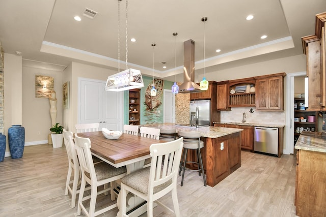 kitchen featuring hanging light fixtures, appliances with stainless steel finishes, a tray ceiling, light stone counters, and island exhaust hood