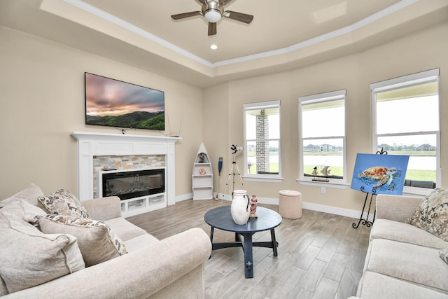 living room featuring a stone fireplace, ceiling fan, light wood-type flooring, ornamental molding, and a tray ceiling