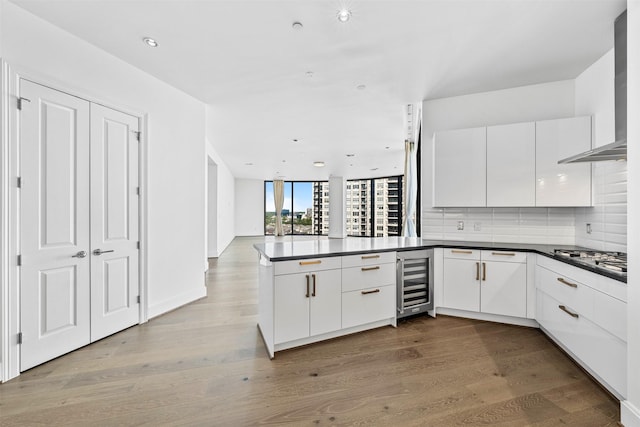 kitchen with wall chimney range hood, wine cooler, kitchen peninsula, stainless steel gas stovetop, and white cabinets