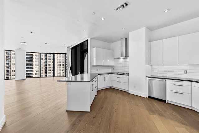 kitchen featuring white cabinets, wall chimney range hood, light hardwood / wood-style flooring, stainless steel dishwasher, and kitchen peninsula