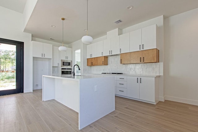 kitchen with backsplash, stainless steel appliances, a kitchen island with sink, pendant lighting, and white cabinets