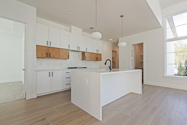 kitchen featuring hanging light fixtures, cooktop, tasteful backsplash, a kitchen island with sink, and white cabinets