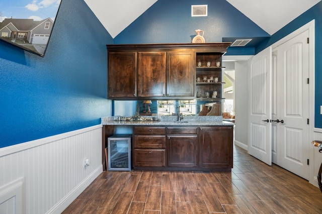 bar with dark brown cabinets, light stone countertops, wine cooler, and vaulted ceiling