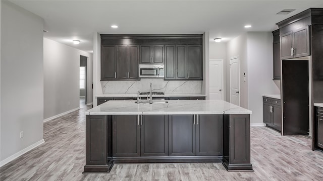 kitchen featuring a kitchen island with sink, sink, light hardwood / wood-style flooring, decorative backsplash, and light stone countertops