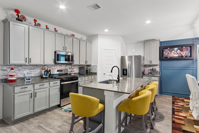 kitchen with dark stone counters, stainless steel appliances, sink, gray cabinets, and an island with sink