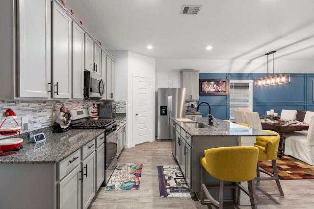 kitchen with gray cabinetry, sink, hanging light fixtures, stainless steel appliances, and an island with sink