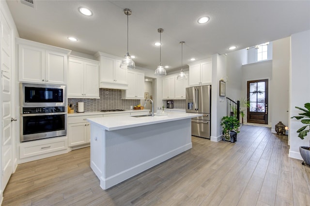 kitchen featuring appliances with stainless steel finishes, light wood-type flooring, a kitchen island with sink, white cabinets, and hanging light fixtures