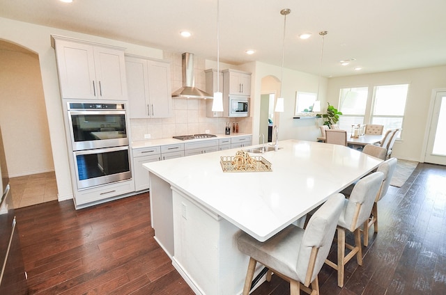 kitchen with a center island with sink, wall chimney range hood, hanging light fixtures, appliances with stainless steel finishes, and white cabinetry